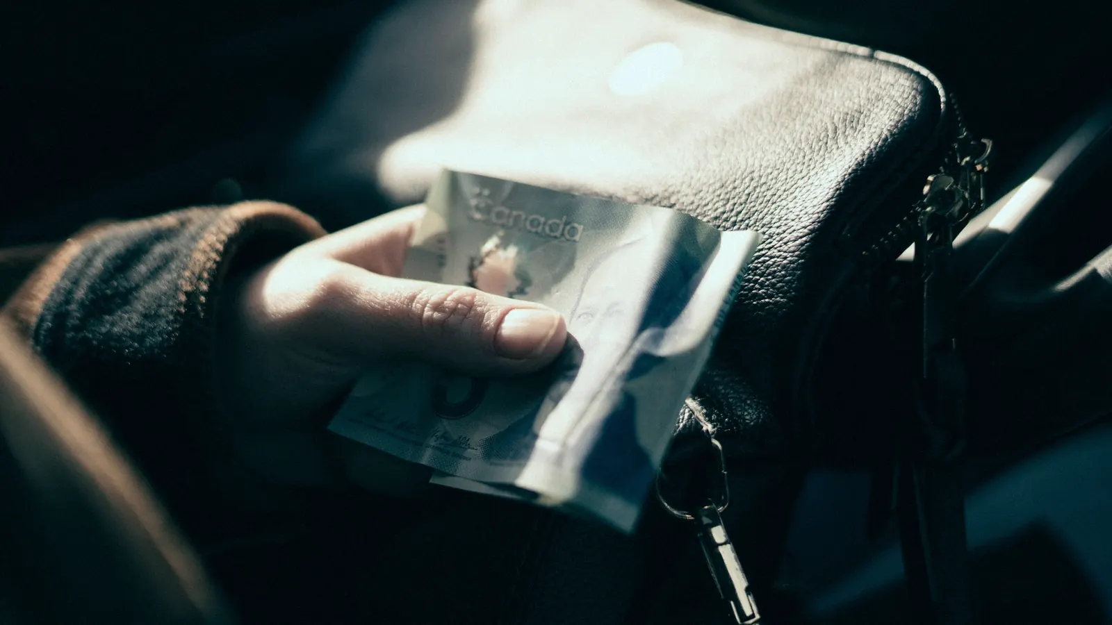 A hand taking a Canadian five-dollar bill out of a leather bag.