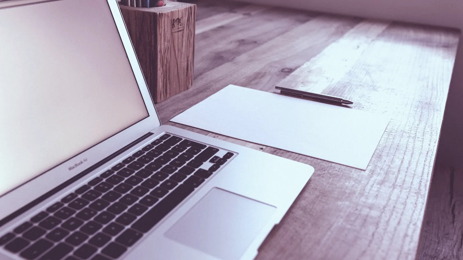 An open laptop on a wooden desk beside a sheet of paper and a pen.