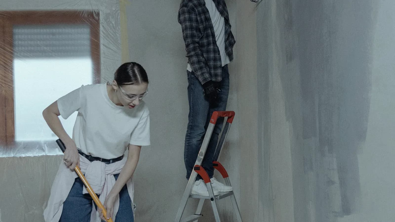 Two people painting the walls of a room under renovation, one on a stepladder.