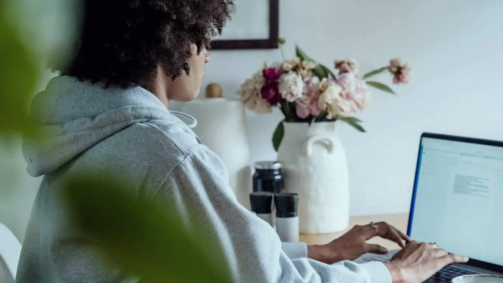 A person working on a laptop at a small desk at home, beside a vase of flowers.