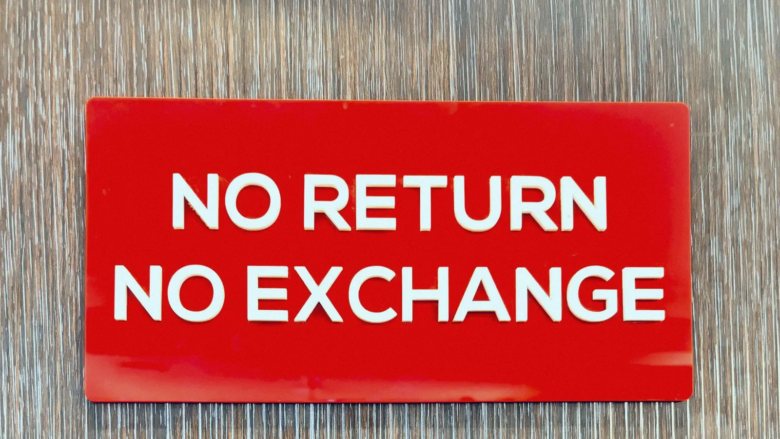 A red plastic shop sign reading NO RETURN NO EXCHANGE mounted on a wooden wall.