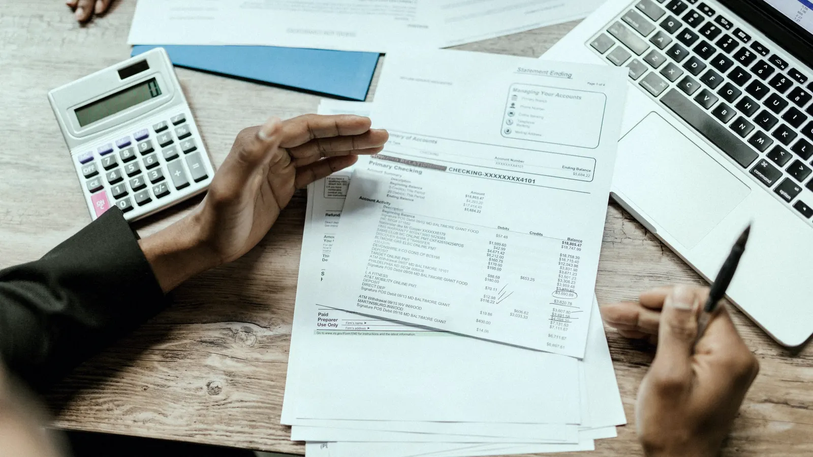 Hands going through a printed statement beside a calculator and a laptop.