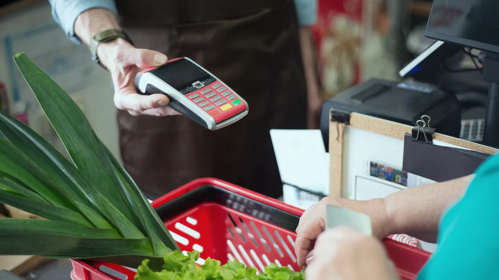 A customer paying by card at a shop counter, with groceries in a basket.