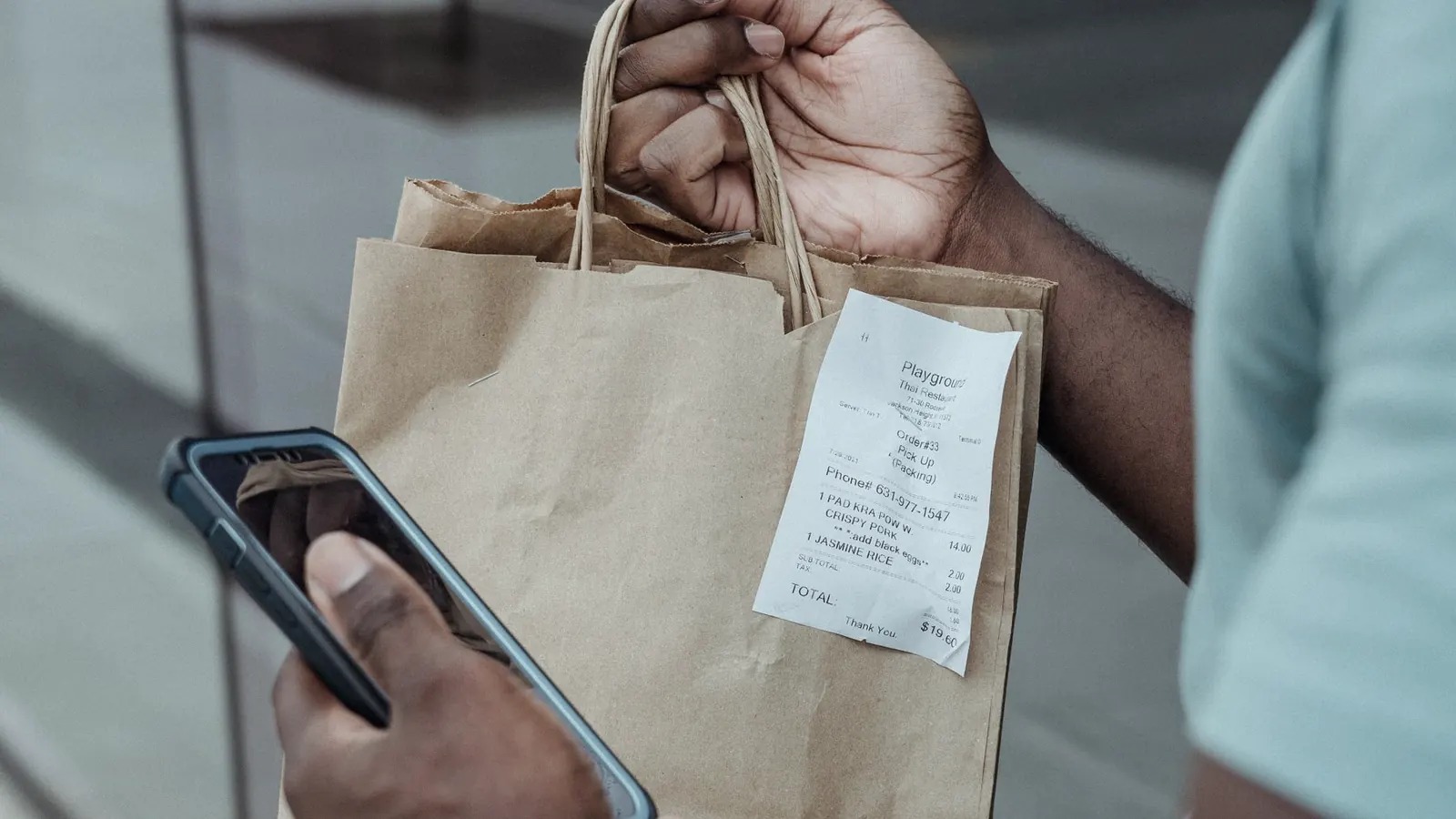 A shopper outside a store holding a takeaway bag with the paper receipt attached, phone in the other hand.