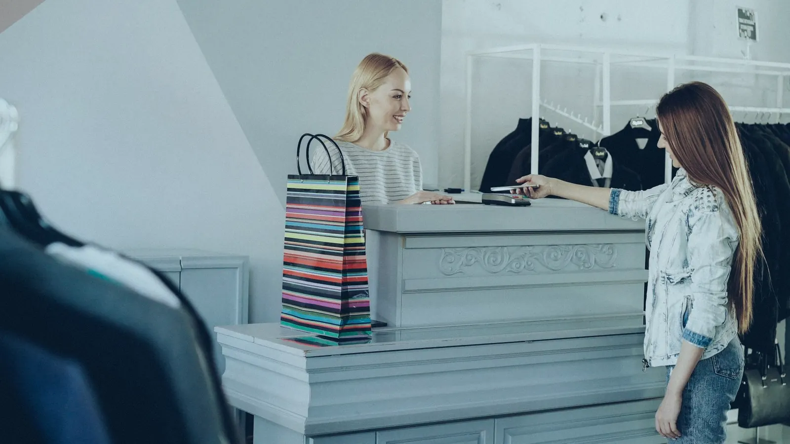 A customer at a clothing shop counter holding out a phone to the staff member serving her.