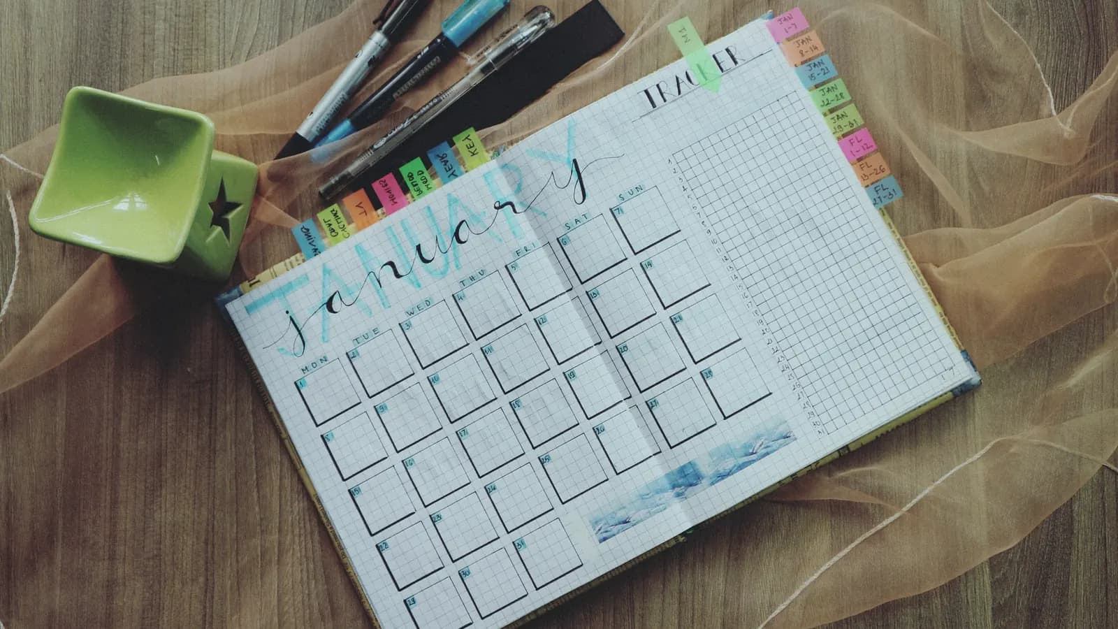 A planner open at a hand-lettered January page, with pens and coloured tabs on a wooden desk.