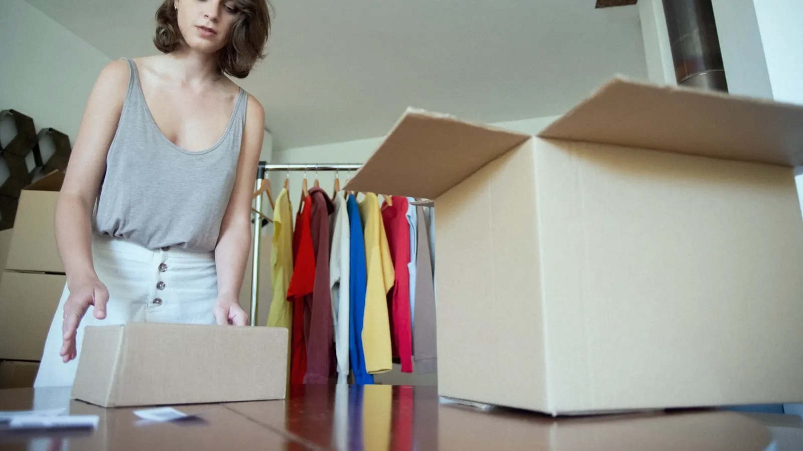 A woman packing orders into boxes in front of a rail of clothes.