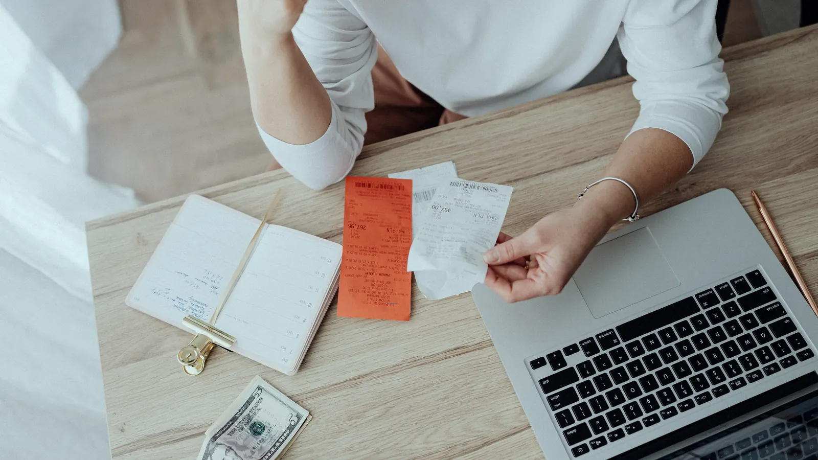A person at a desk going through paper receipts beside a laptop, with a notebook and a dollar bill.