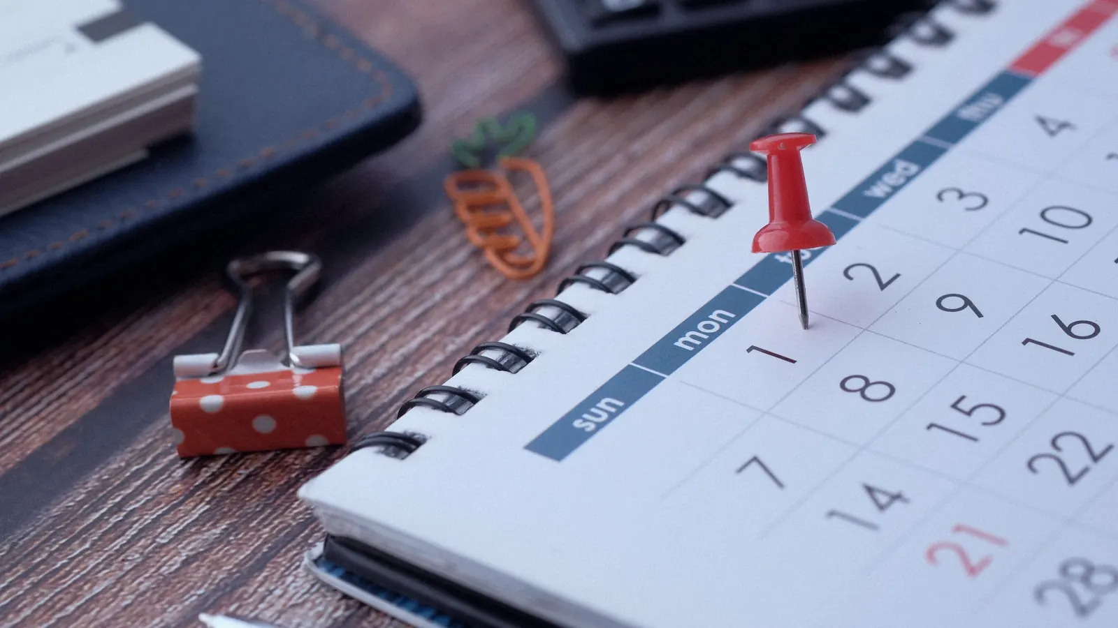 A red pin marking a date on a desk calendar, beside a pen and binder clips.