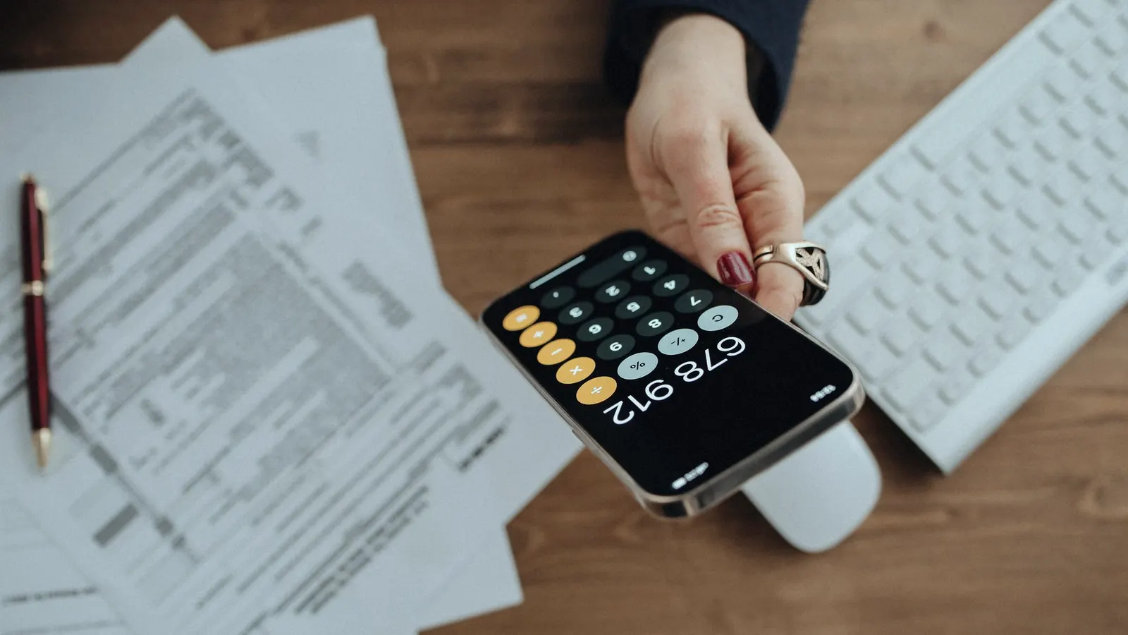 A hand holding a smartphone over printed tax records, beside a pen and a keyboard.