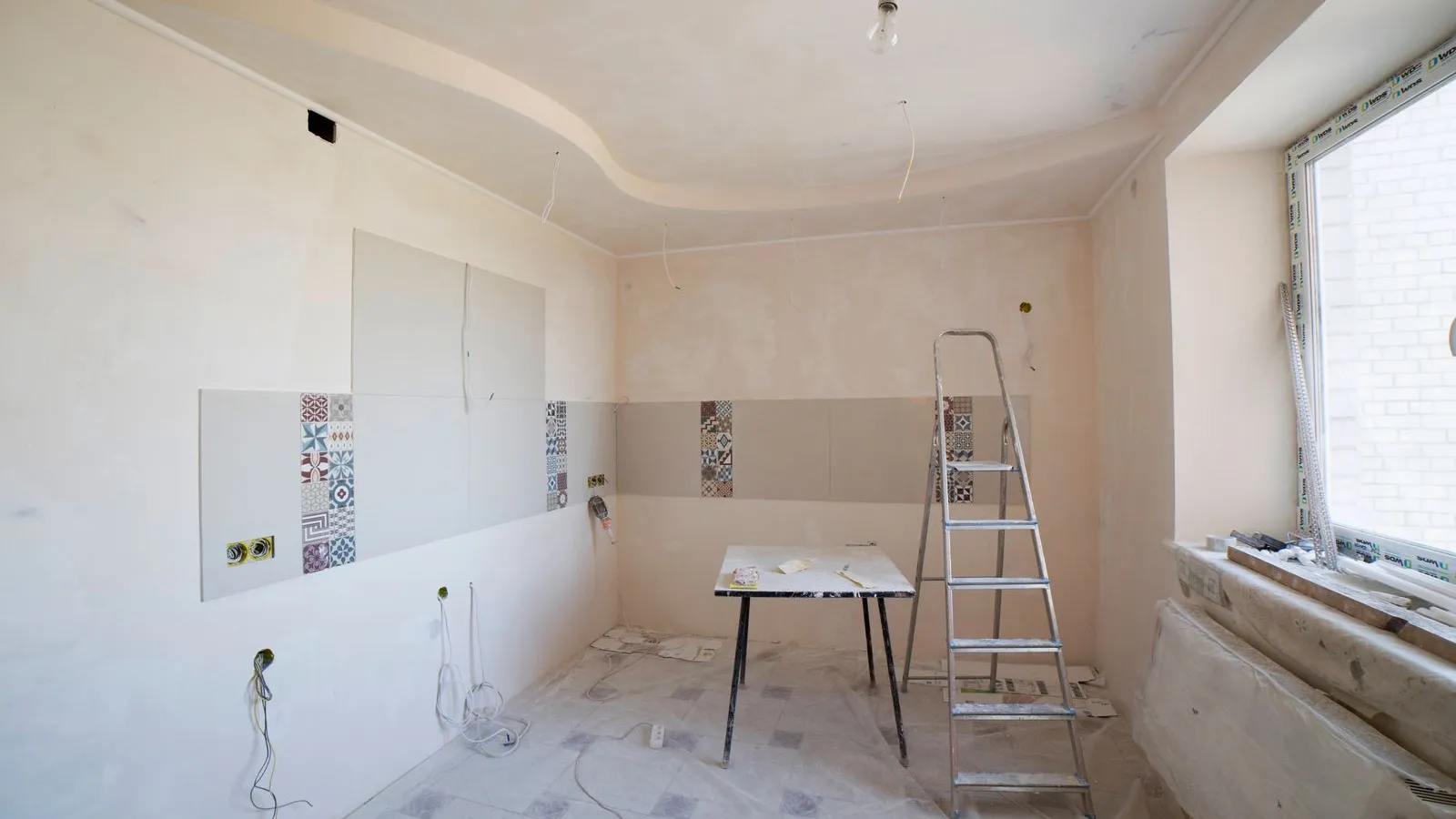 A kitchen mid-renovation with bare plastered walls, sample tiles, a stepladder and a trestle table.